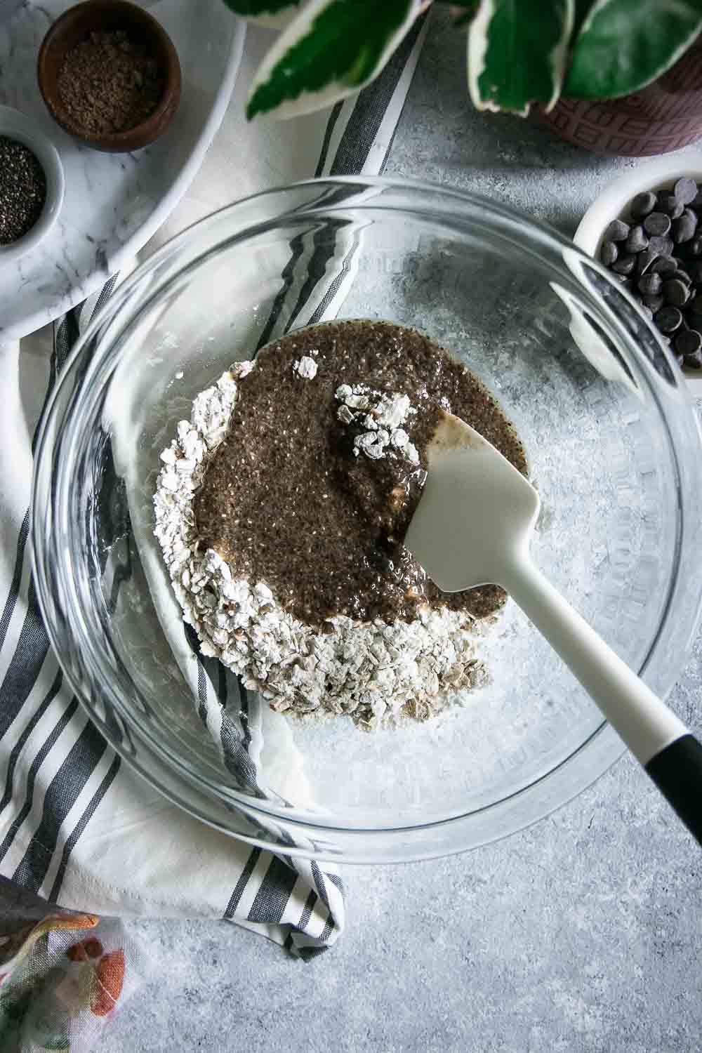 a glass bowl with dry and wet oatmeal chocolate chip cookies and a mixing spoon on a grey table