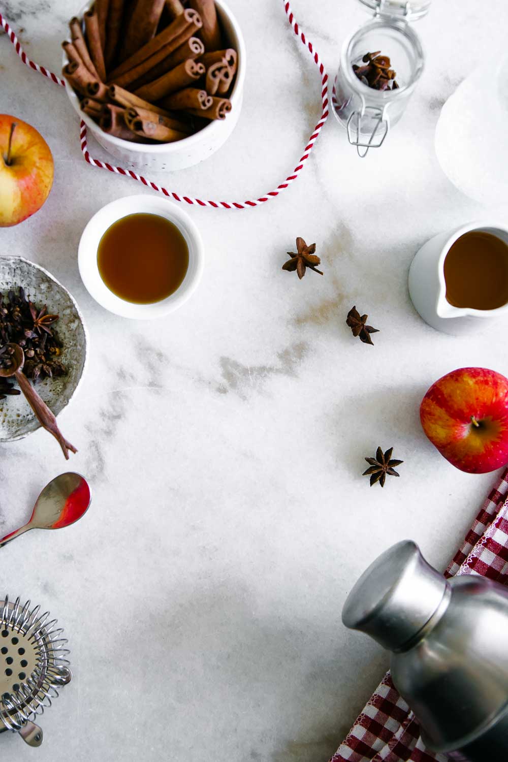 ingredients for a fall spritzer on a white marble countertop, including apples, sparkling water, maple syrup, cloves, anise, and cinnamon