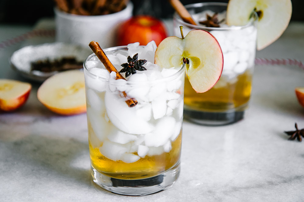 an apple cider vinegar spritzer over ice in a cocktail glass with an apple slice and cinnamon stick garnish on a white table