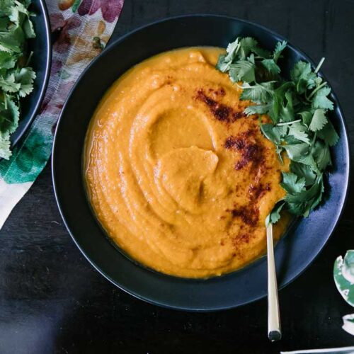 A bowl of orange soup made with sweet potatoes, carrots, and spicy chipotle with cilantro on top on a dark table
