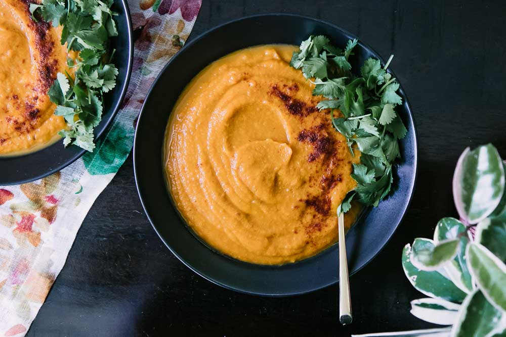 A bowl of orange soup made with sweet potatoes, carrots, and spicy chipotle with cilantro on top on a dark table