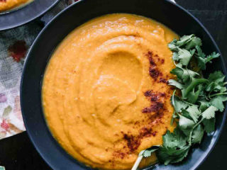 a bowl of sweet potato and carrot soup with cilantro as garnish on a wood table with a gold spoon with the words "sweet potato carrot soup" in black and white writing