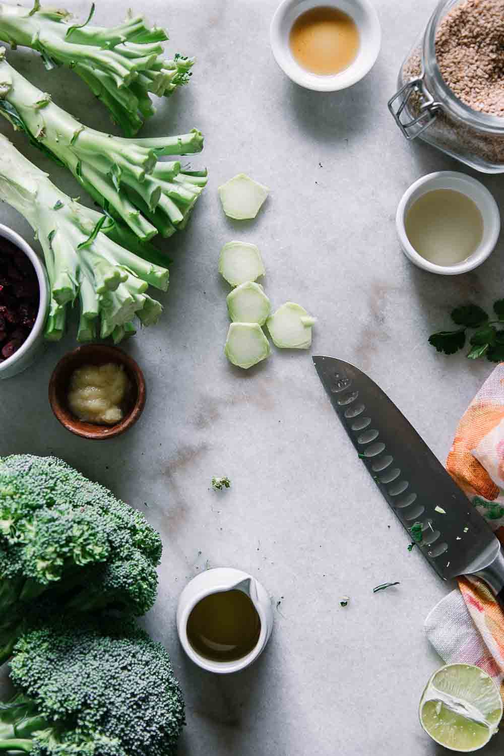 a cutting board with a knife, cut broccoli stalks, ginger, oil, and a lime