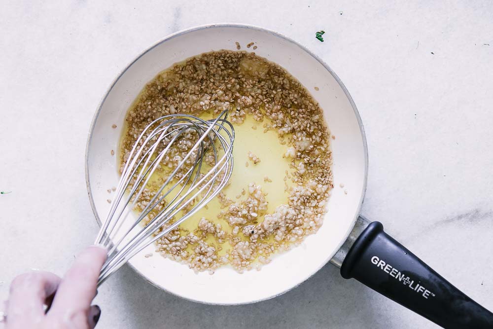 a white saucepan with toasted sesames, garlic, oil, and a hand whisk