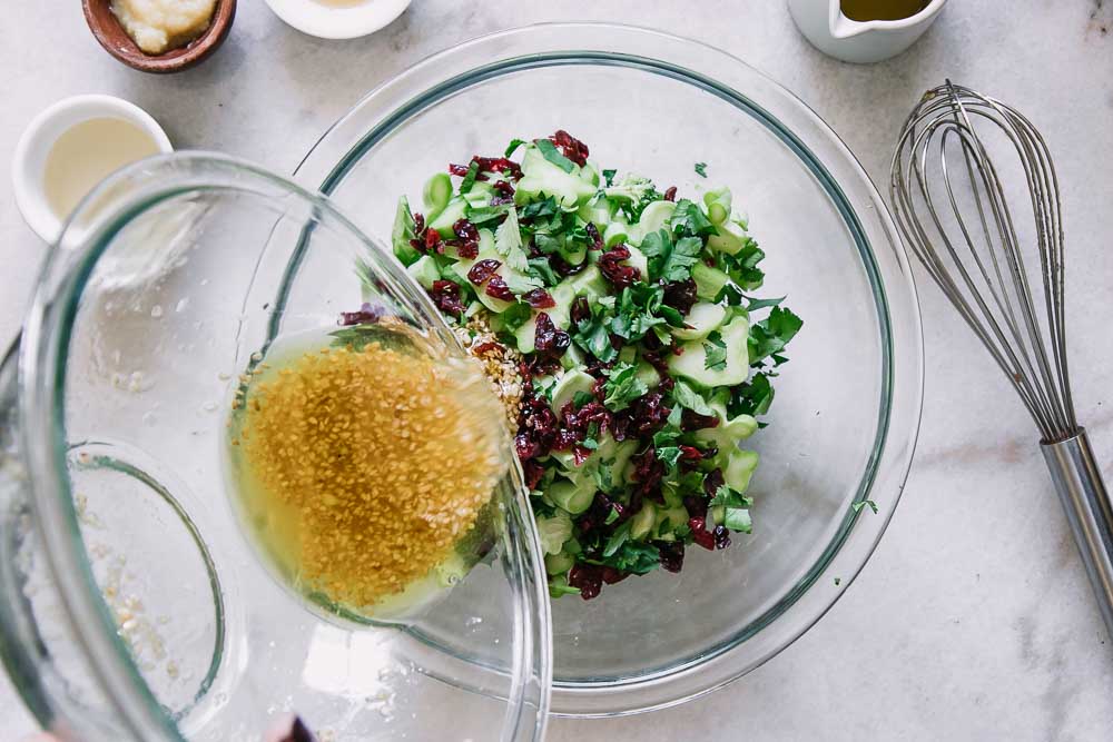 a hand pouring a bowl of salad dressing into a green broccoli and cranberry salad