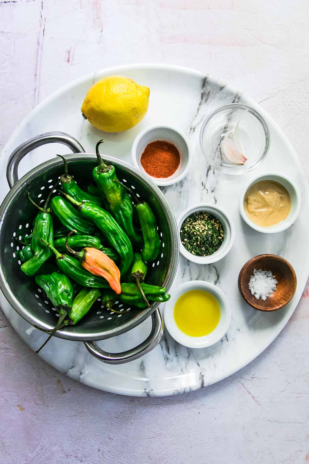 A round white plate with green shishito peppers and small bowls of oil, spices, and a lemon