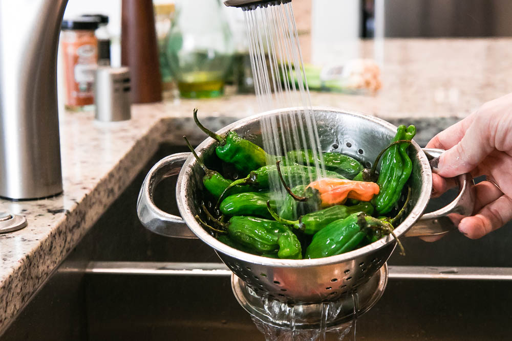 a hand washing green shishito peppers in a metal colander under running water