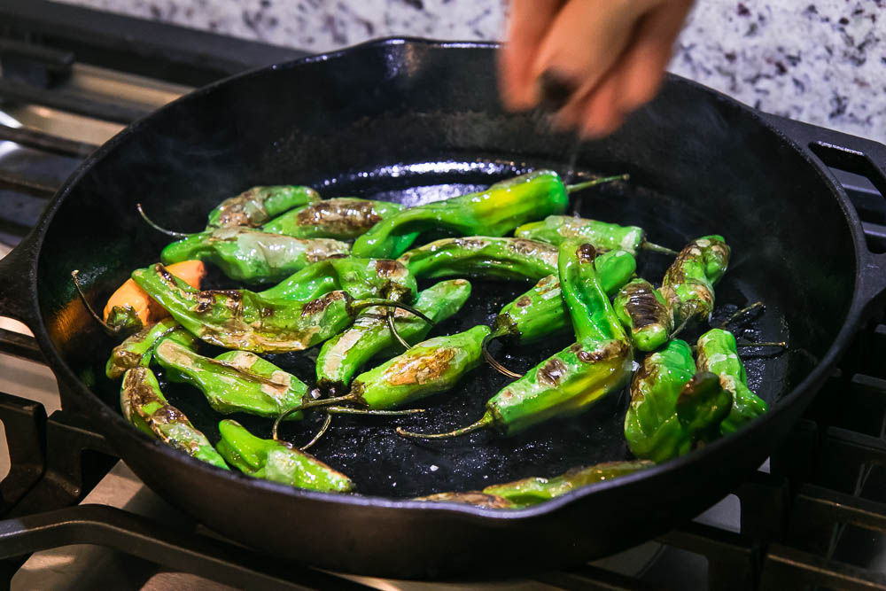 a hand sprinkling salt onto shishito peppers cooking in a cast iron skillet