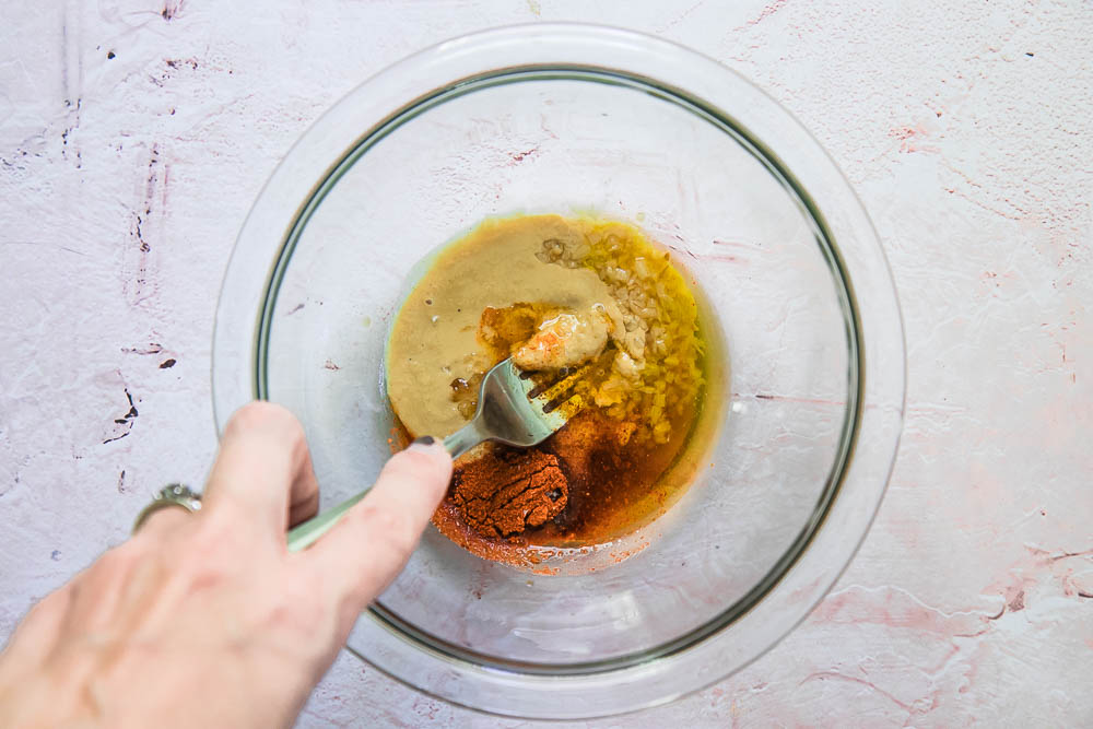 a hand mixing a sauce together with a fork in a glass mixing dish on a white table