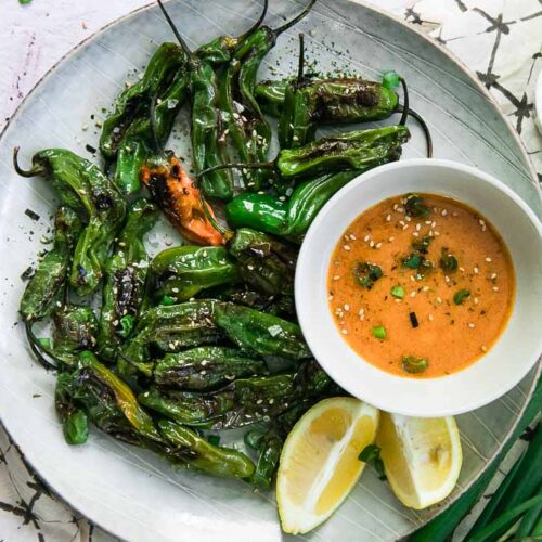 a white plate with cooked small green peppers and an orange dipping sauce