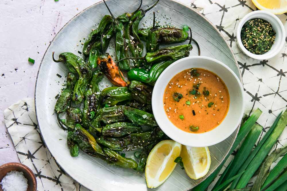 a white plate with cooked small green peppers and an orange dipping sauce