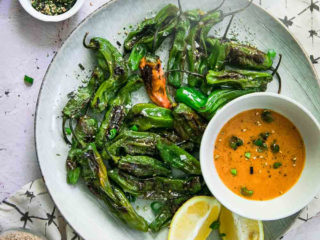 a plate of cooked shishito peppers with an orange dipping sauce and the words "pan roasted shishito peppers" in orange and black writing