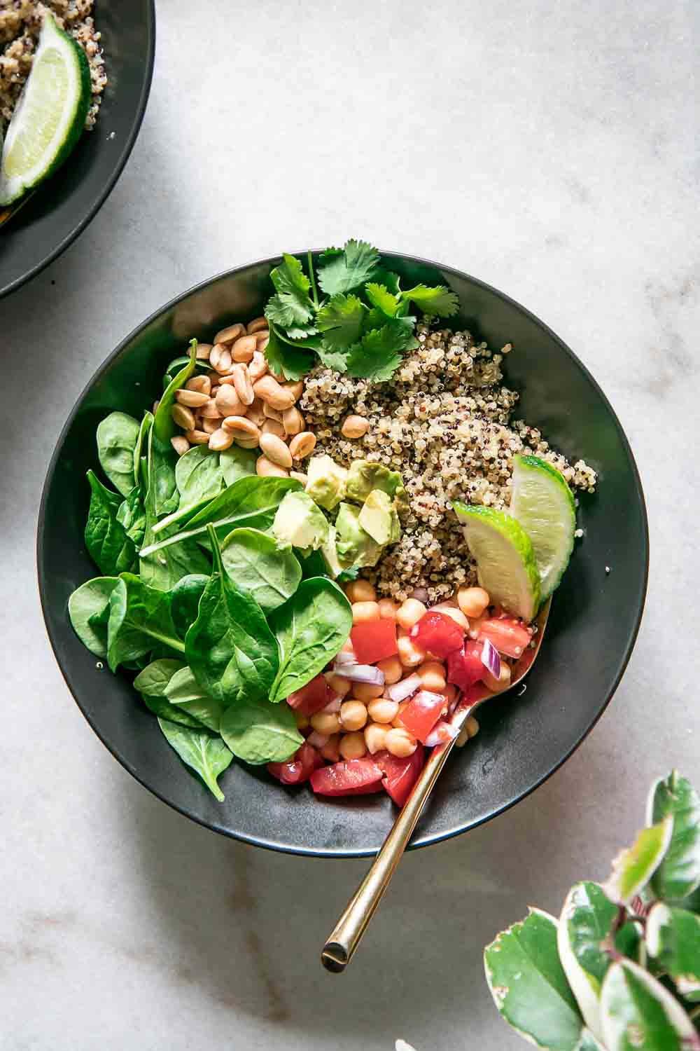 a black bowl with quinoa, spinach, chickpeas, tomatoes, and limes with a gold spoon