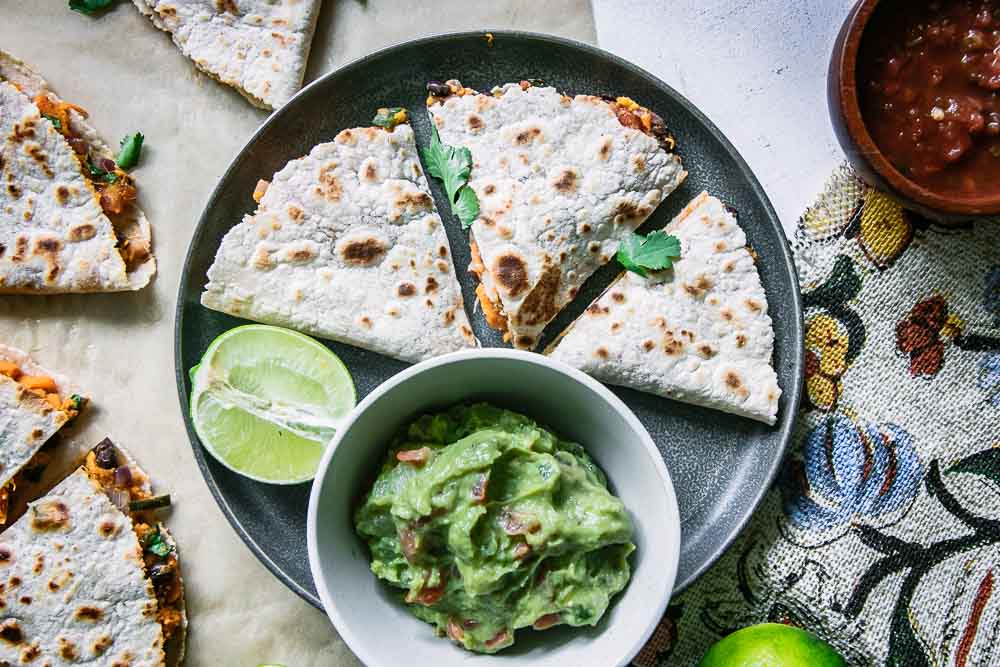 a blue plate with a cut vegan quesadilla with a bowl of guacamole and a lime wedge