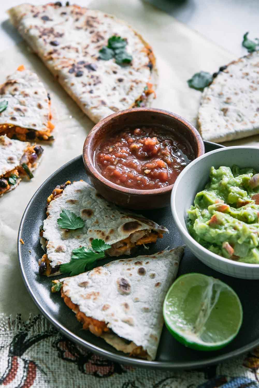 a plant-based quesadilla on a plate with cut quesadilla pieces on a table with small bowls of guacamole and salsa