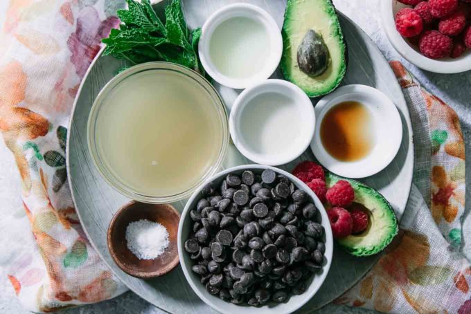 a bowl of chocolate chips, a bowl of aquafaba, vanilla, lemon juice, salt, avocado, and raspberries on a white plate on a blue table