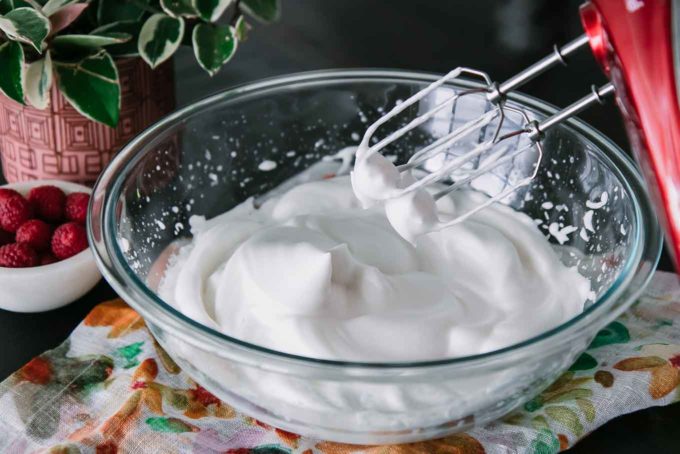 a bowl of aquafaba with stiff peaks and a handheld mixer on a wooden table