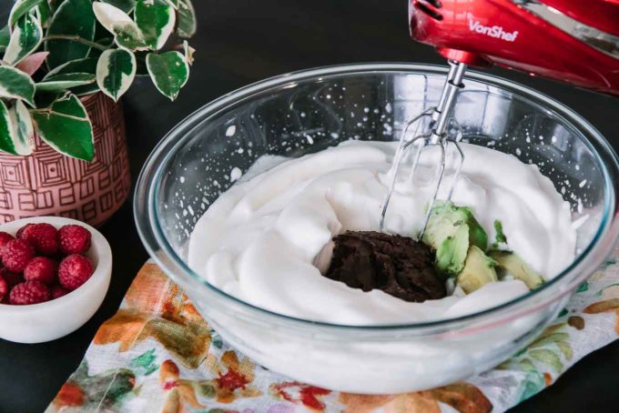 a mixing bowl with aquafaba, melted chocoalte, avocado, and a red handheld electric mixer