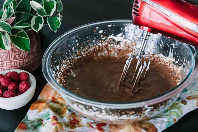 a glass mixing bowl with blended chocolate and a red handheld electric mixer on a wood table
