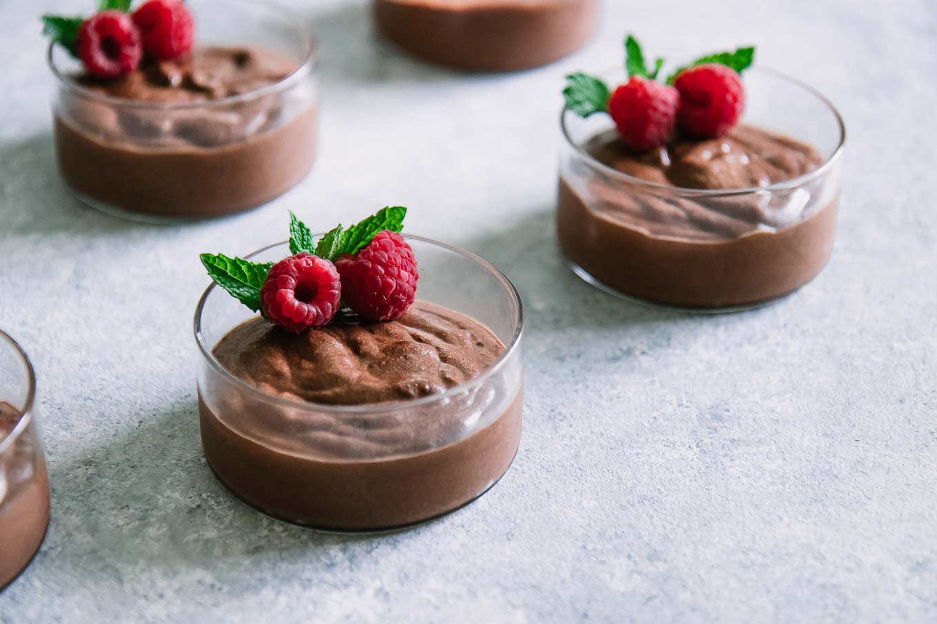small round glass bowls of chocolate mousse with raspberries and mint on top of a blue table