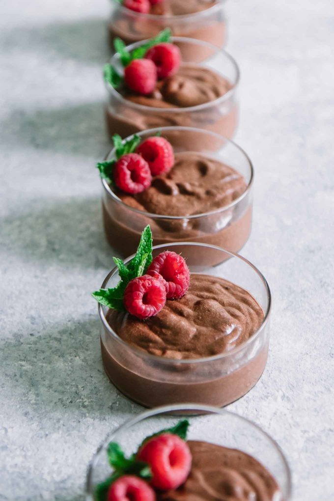 small round glass ramekins of chocolate mousse with raspberries and mint lined up in a row on a blue table