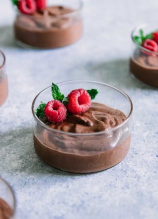a glass bowl with brown chocolate mousse, red raspberries, and a sprig of green mint on a blue table