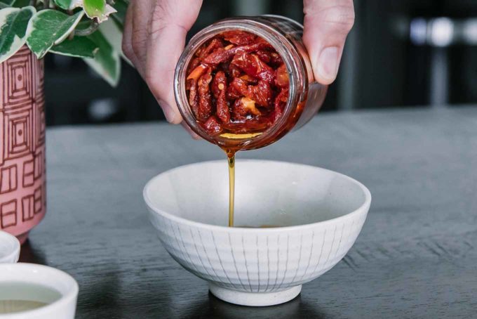 a hand pouring oil from a jar of sun-dried tomatoes into a small white bowl