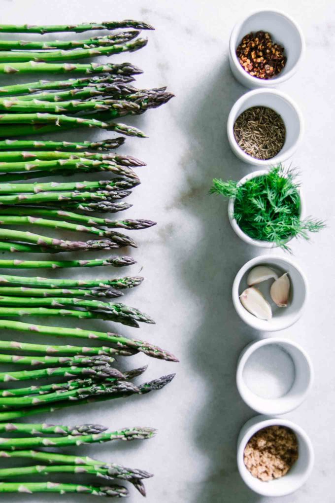 asparagus spears lined up on a white counter with bowls of salt, sugar, dill, garlic, and caraway seeds