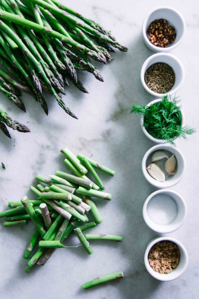 cut asparagus stems and stalks on a white countertop with bowls of herbs, salt, sugar, and spices