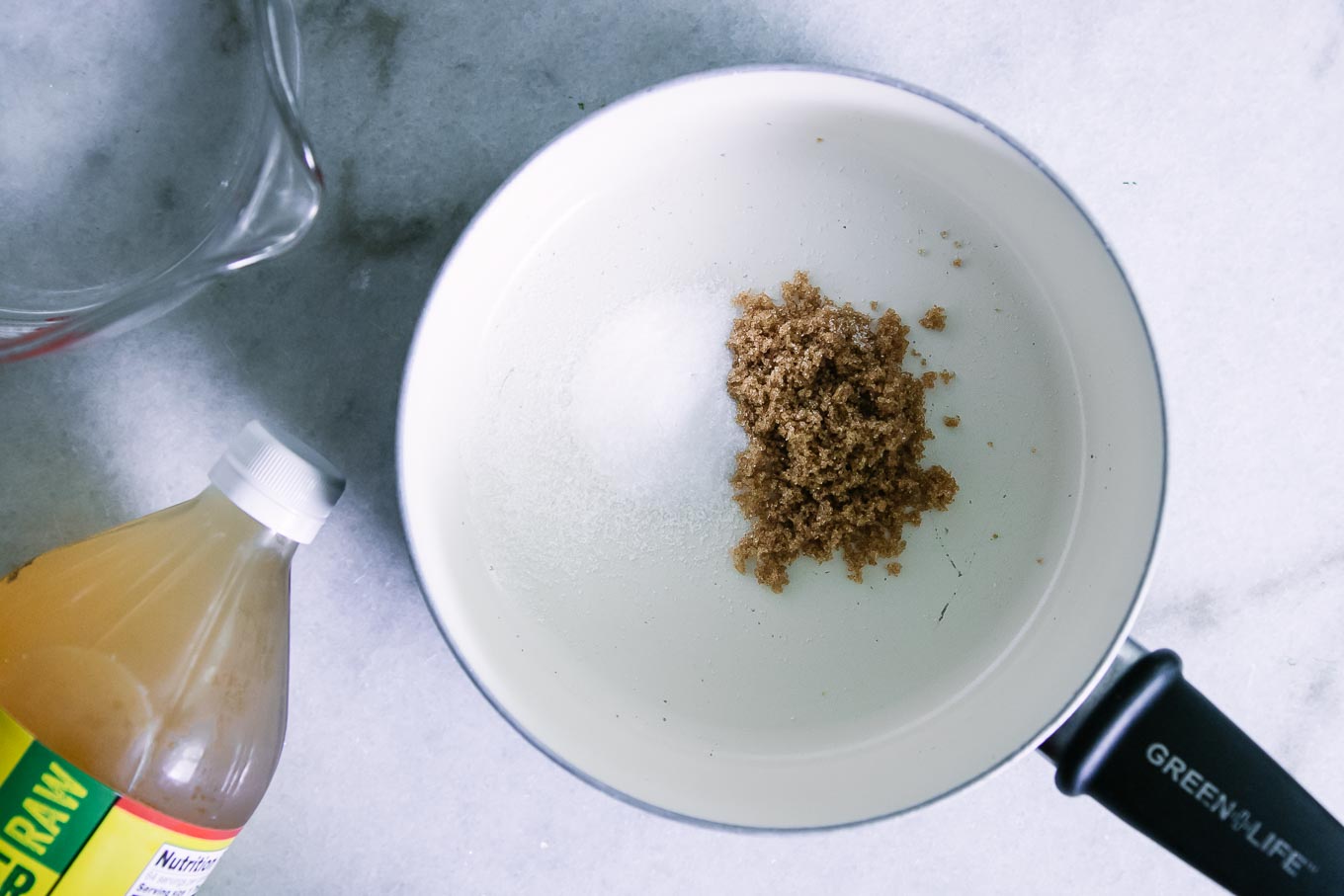 a small sauce pan with salt and sugar inside on a white table with a measuring glass of water and a bottle of apple cider vinegar