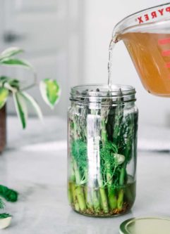 apple cider vinegar brine pouring into a tall glass jar of asparagus with herbs and spices on a white table