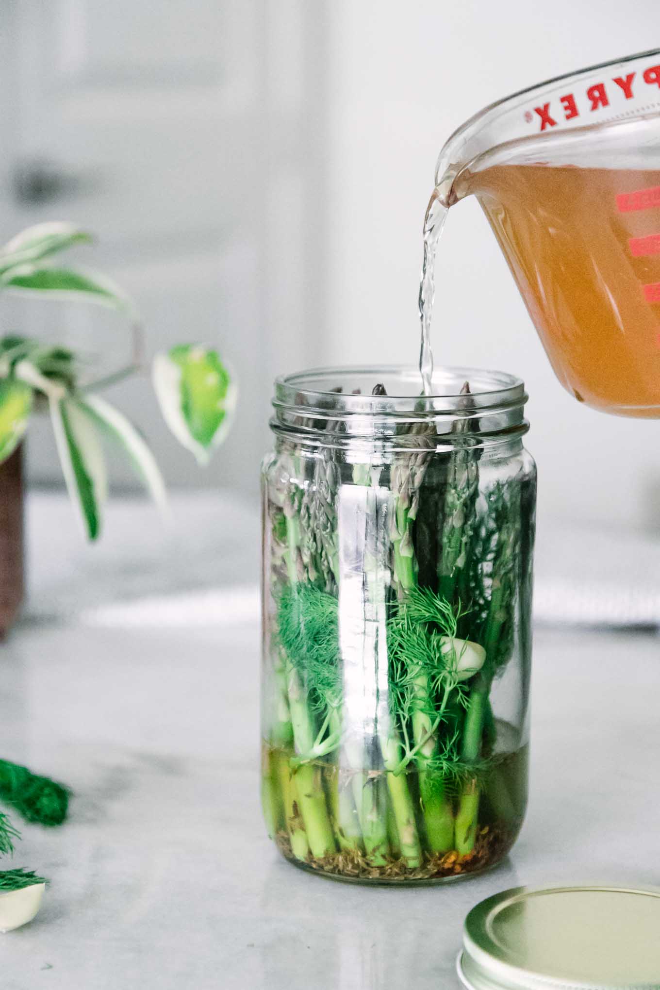 apple cider vinegar brine pouring into a tall glass jar of asparagus with herbs and spices on a white table