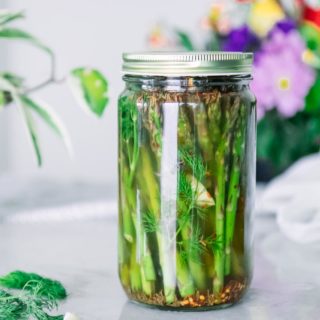 a glass jar filled with asparagus, apple cider vinegar brine, dill, and garlic on a white table