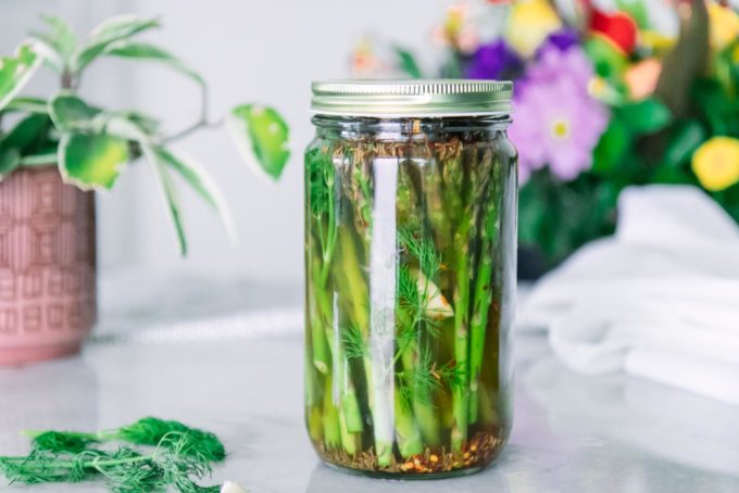 a glass jar filled with asparagus, apple cider vinegar brine, dill, and garlic on a white table