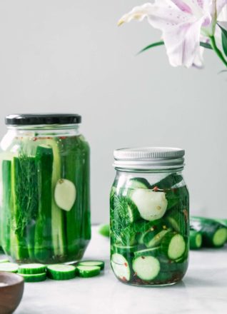 a jar of sliced pickles and a jar of pickle spears on a white table with a pink flower