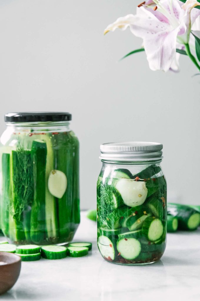 a jar of sliced pickles and a jar of pickle spears on a white table with a pink flower