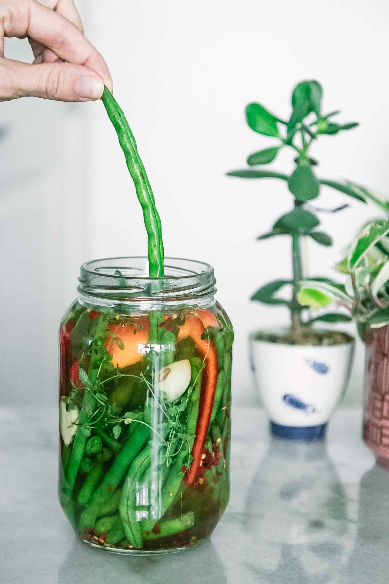 a hand pulling a green bean out of a jar on a white table