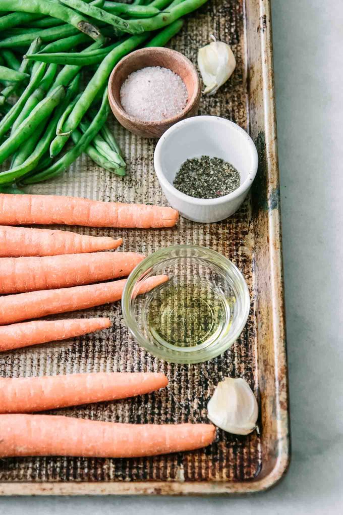 a sheet pan with carrots, green beans, garlic cloves, and bowls of salt and black pepper