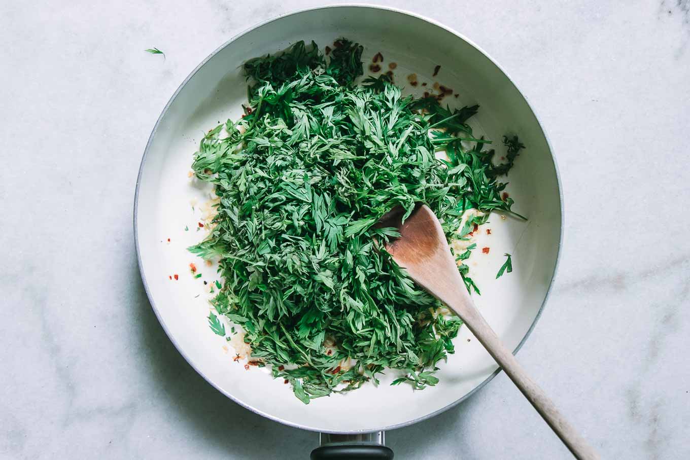 a white pan with carrot leaves and oil and a wooden spoon