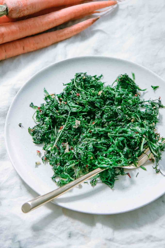 sauteed carrot greens on a white plate on a white table with carrots in the background