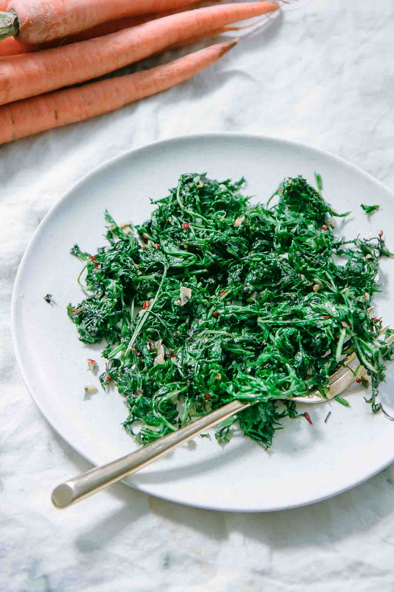 sauteed carrot greens on a white plate on a white table with carrots in the background