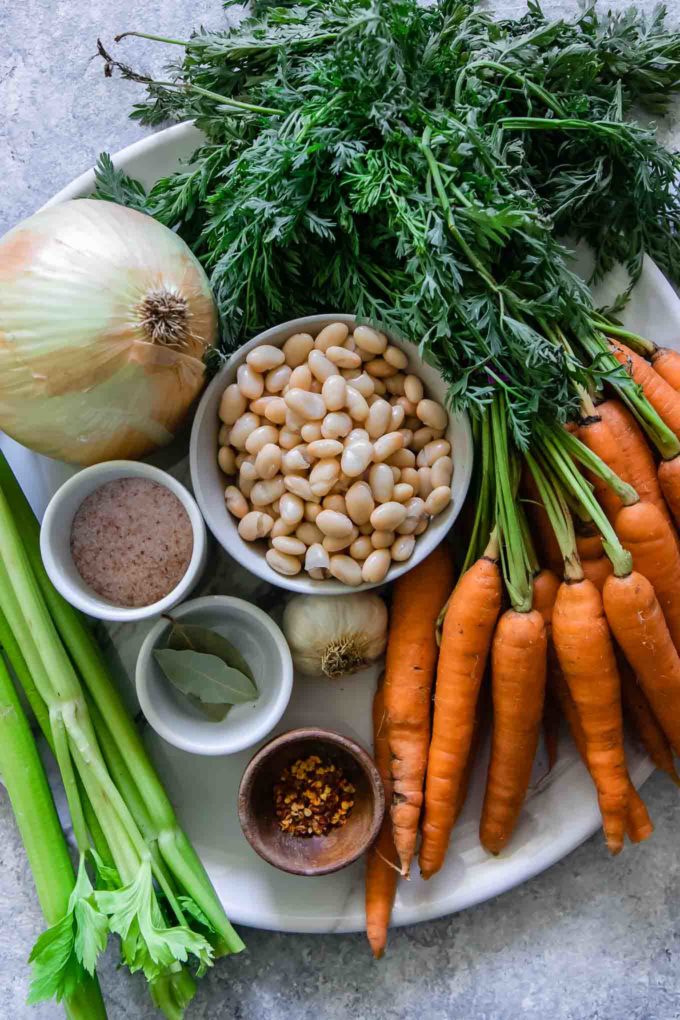 carrots with greens tops, onion, a bowl of white beans, celery, and spices on a round white plate