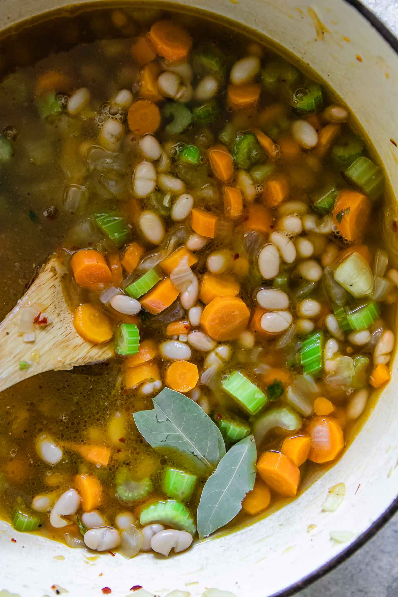 a wooden spoon stirring vegetable soup with carrots and bay laves in a large soup pot