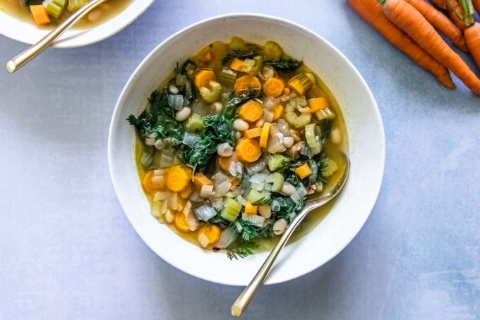 a bowl of vegetable soup with leafy carrot greens in a white bowl on a blue table