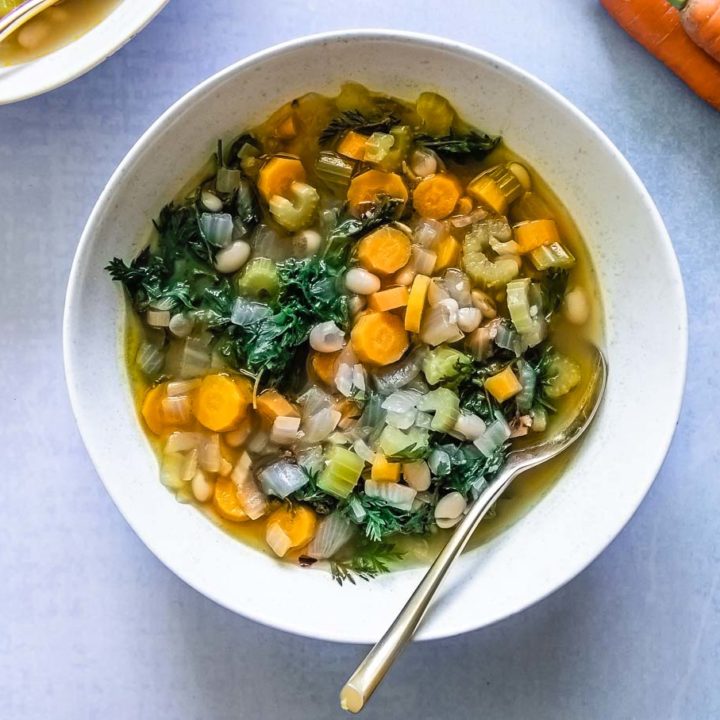 a bowl of vegetable soup with leafy carrot greens in a white bowl on a blue table