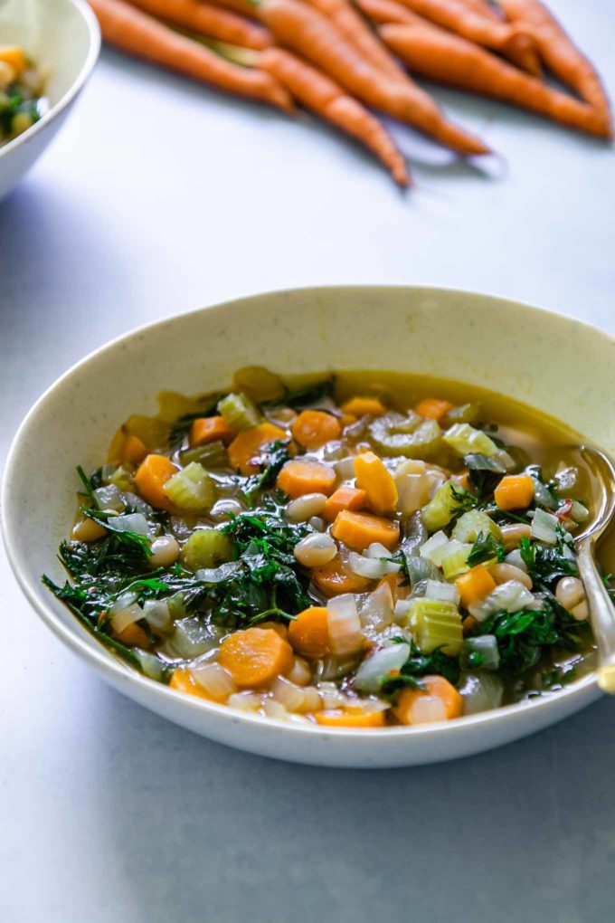 a white bowl of soup with sliced carrots, carrot leaves, onion, and beans on a blue table with carrots in the background