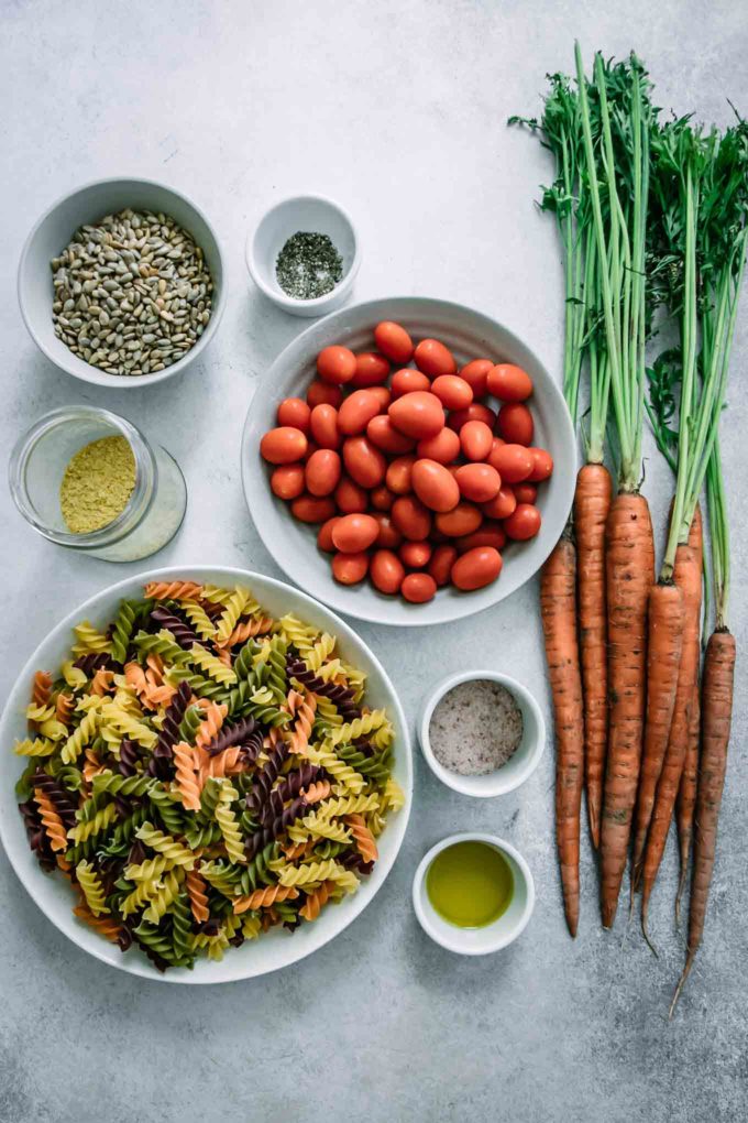 bowls of dry rotini pasta, cherry tomatoes, carrots with tops, oil, salt, sunflower seeds, cheese, and pepper on a white table