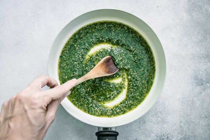 a hand stirring green carrot leaves pesto in a white pan