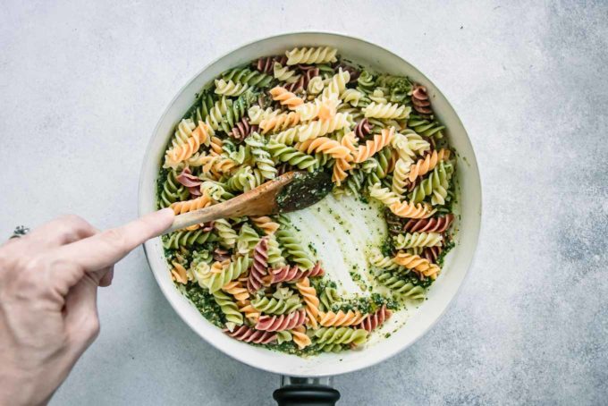 a hand stirring pesto pasta in a white pan with a wooden spoon