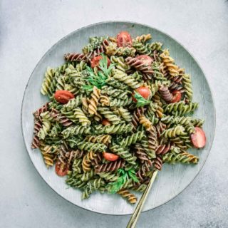 pesto pasta with cherry tomatoes on a white plate on a white table with carrots and a bowl of tomatoes